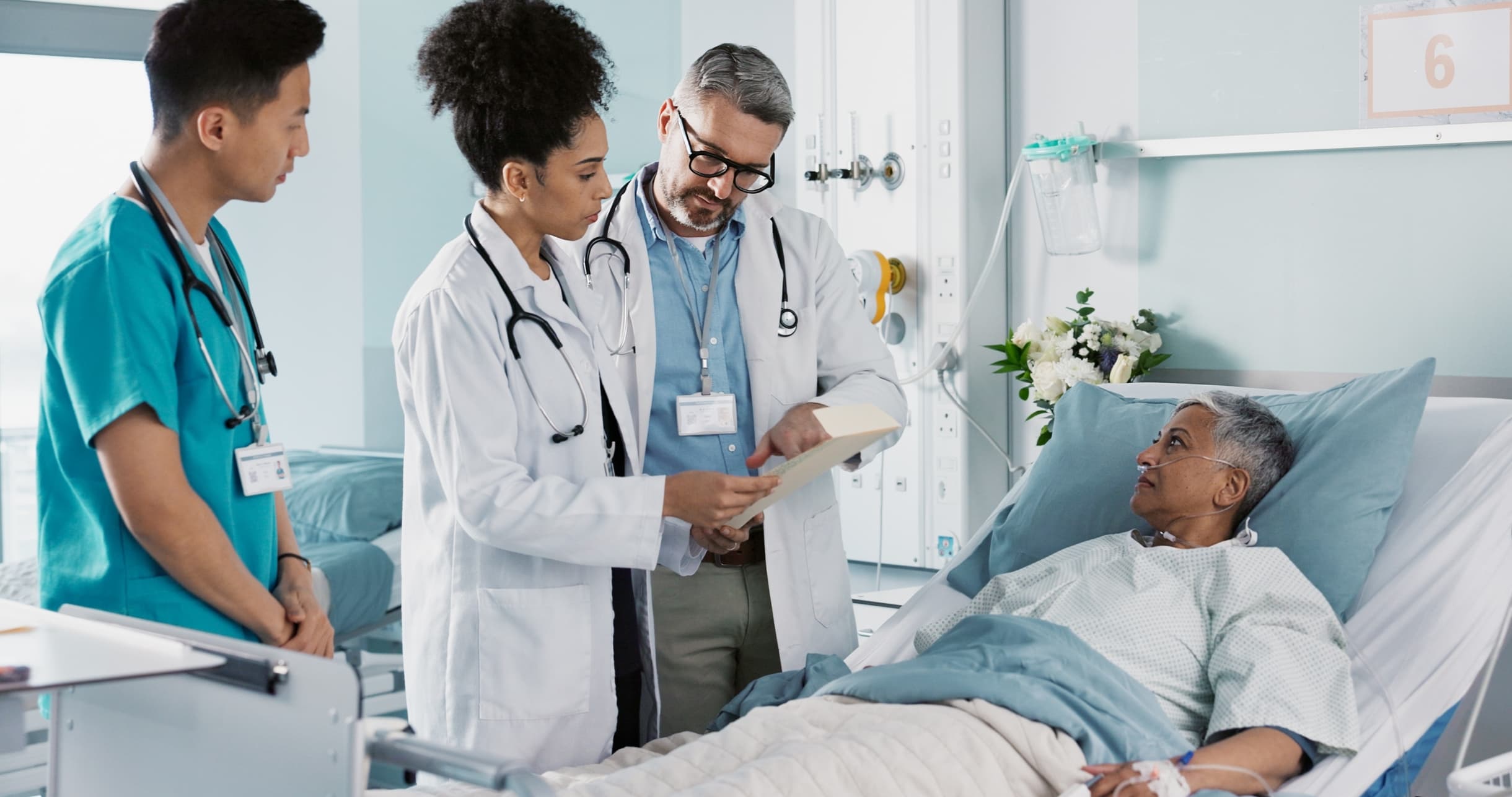 Three doctors review a patient's chart as the patient lays in bed listening