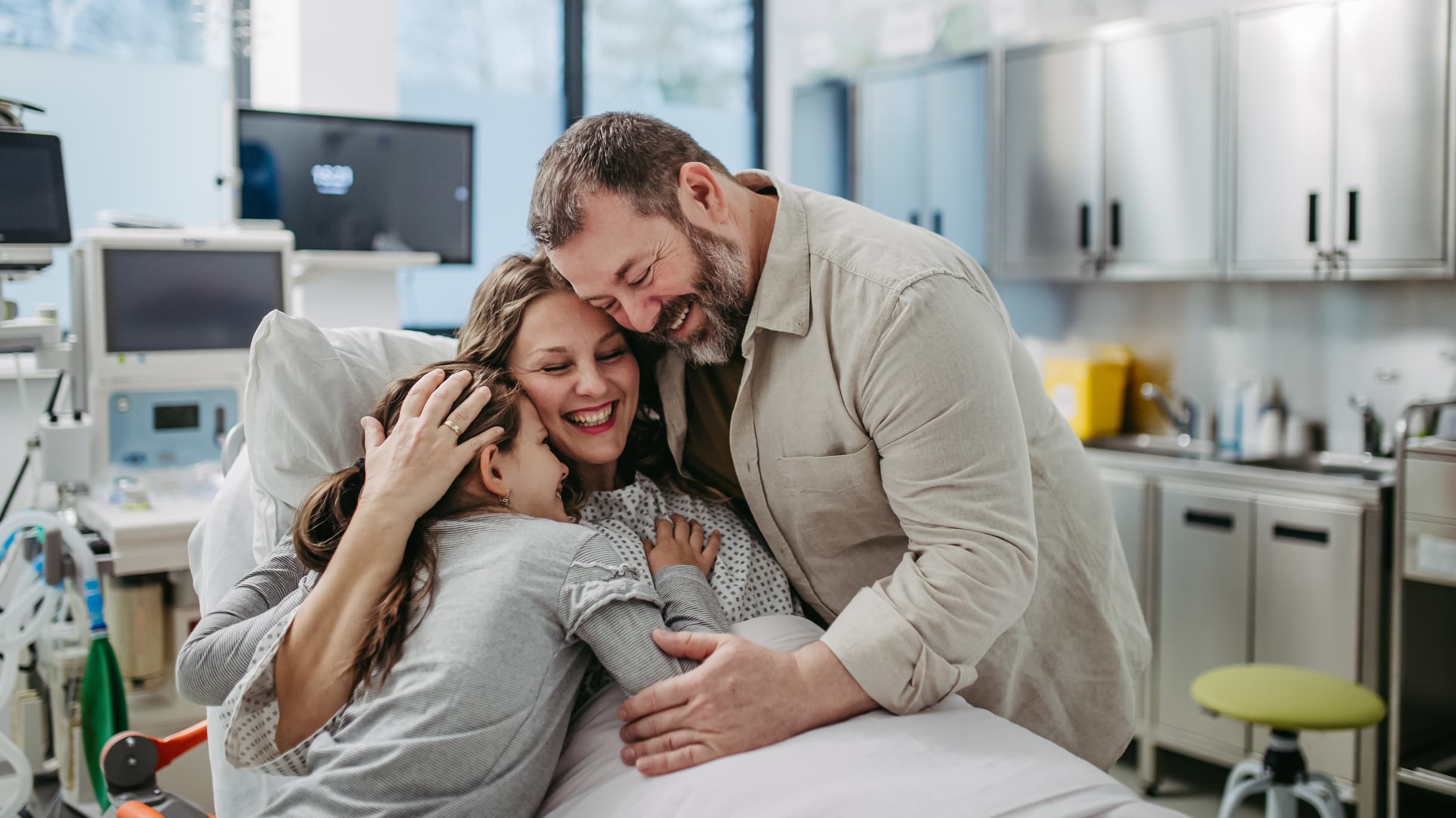 A husband and daughter hug a woman in a hospital bed